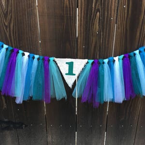 Puede incluir: Una pancarta triangular blanca con el n&uacute;mero "1" en verde. La pancarta est&aacute; enhebrada con serpentinas de tul azul, morado y blanco.