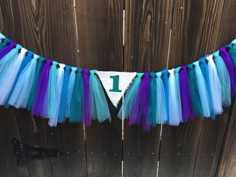 Puede incluir: Una pancarta triangular blanca con el n&uacute;mero "1" en verde. La pancarta est&aacute; enhebrada con serpentinas de tul azul, morado y blanco.