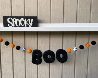 Pode incluir: Uma faixa de feltro preto com a palavra "BOO" em letras brancas, pendurada em uma prateleira branca com uma placa preta que diz "Spooky" acima. A faixa é decorada com bolas de feltro laranja e preto.