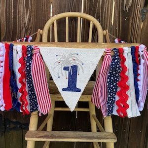 May include: A white burlap banner with a blue number one and silver glitter fireworks. The banner is decorated with red, white, and blue ribbon streamers and pom-poms.