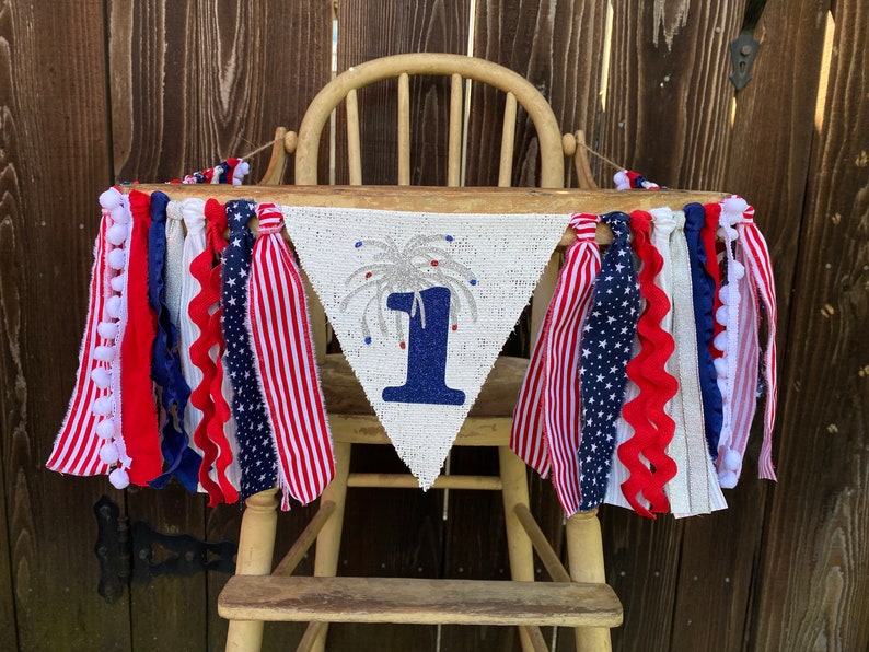 May include: A white burlap banner with a blue number one and silver glitter fireworks. The banner is decorated with red, white, and blue ribbon streamers and pom-poms.