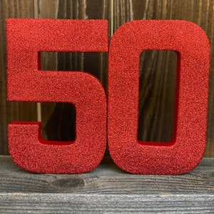 May include: Red glitter number 50, the numbers are standing upright on a wooden surface.