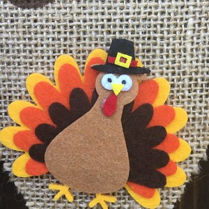 May include: A felt turkey decoration with a black pilgrim hat, yellow feet, and a red wattle. The turkey has a brown body and a colorful fan of feathers in shades of orange, yellow, and brown. The background is a woven burlap material.