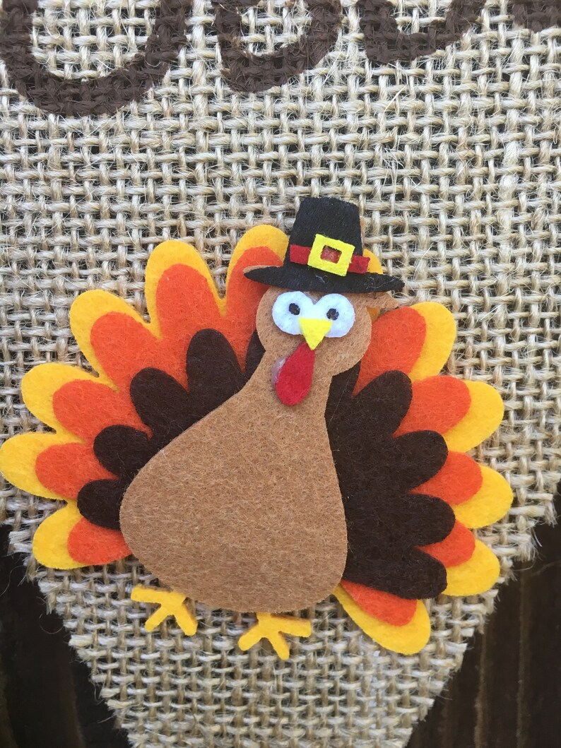 May include: A felt turkey decoration with a black pilgrim hat, yellow feet, and a red wattle. The turkey has a brown body and a colorful fan of feathers in shades of orange, yellow, and brown. The background is a woven burlap material.