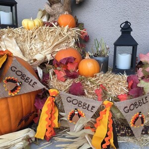 May include: Thanksgiving-themed display featuring pumpkins, hay, and decorative banners. The banners read "GOBBLE" and feature turkey designs. The scene includes lanterns and fall foliage, creating a festive autumn atmosphere.