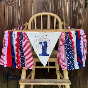 May include: A white burlap banner with a blue number one and a silver glitter firework design. The banner is decorated with red, white, and blue ribbon and pom-poms. The banner is hanging on a wooden high chair.