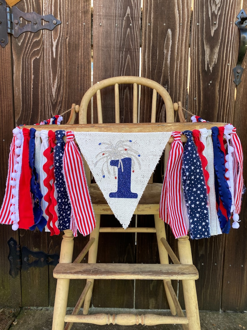 May include: A white burlap banner with a blue number one and a silver glitter firework design. The banner is decorated with red, white, and blue ribbon and pom-poms. The banner is hanging on a wooden high chair.