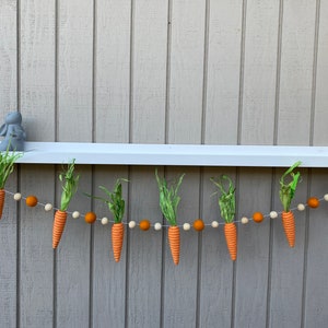 May include: A garland of orange felt carrots with green felt leaves hangs from a white shelf. The garland is strung with orange and white felt balls.