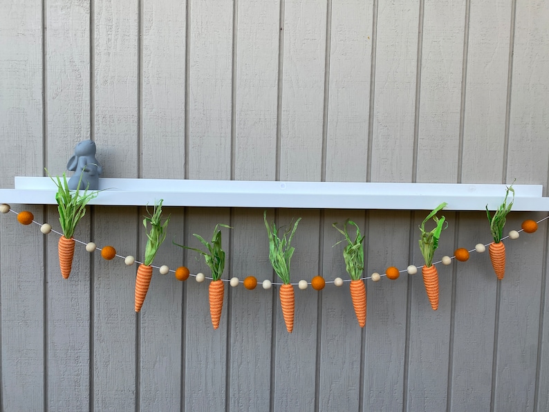 May include: A garland of orange felt carrots with green felt leaves hangs from a white shelf. The garland is strung with orange and white felt balls.