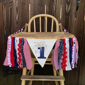 May include: A burlap banner with a blue number one and a firework design, decorated with red, white, and blue fabric streamers. The banner is hanging on a wooden high chair.