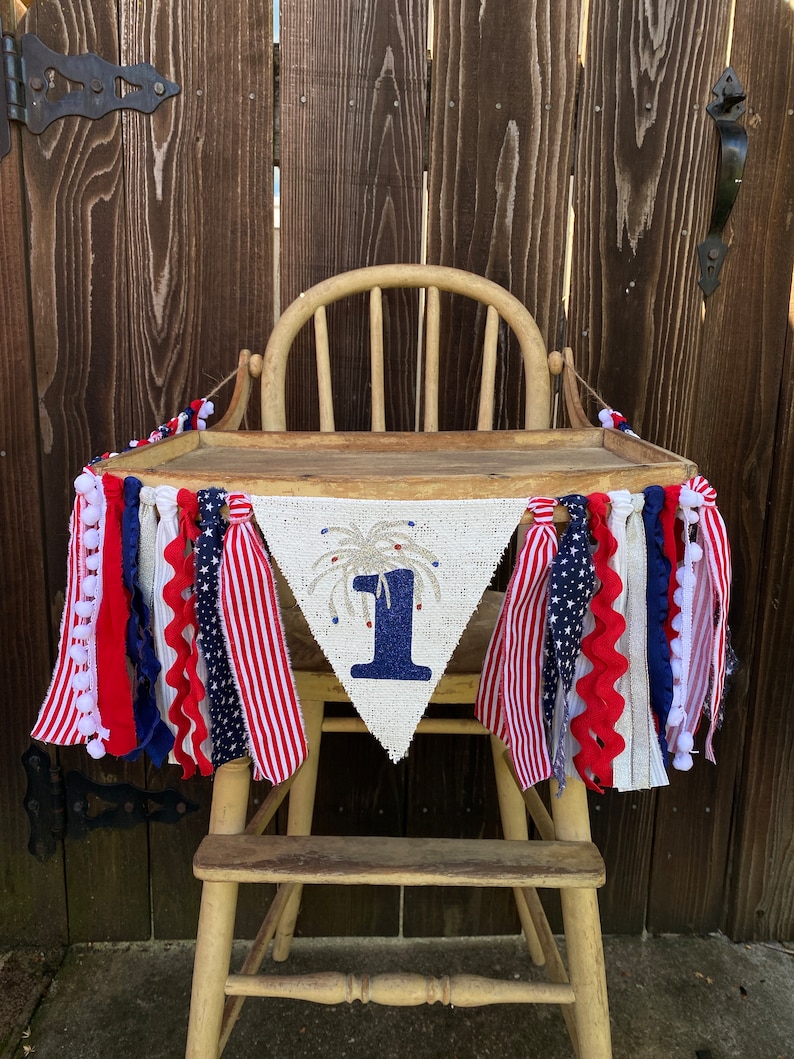 May include: A burlap banner with a blue number one and a firework design, decorated with red, white, and blue fabric streamers. The banner is hanging on a wooden high chair.
