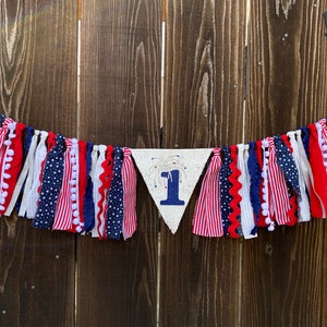 May include: A red, white, and blue fabric banner with a white triangle in the center that says "1" with a firework design. The banner is made of various fabrics with pom-poms and stars.