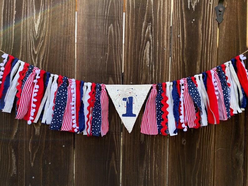 May include: A red, white, and blue fabric banner with a white triangle in the center that says "1" with a firework design. The banner is made of various fabrics with pom-poms and stars.