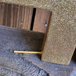 May include: A gold glitter letter with a wooden base. The letter is standing on a wooden surface.