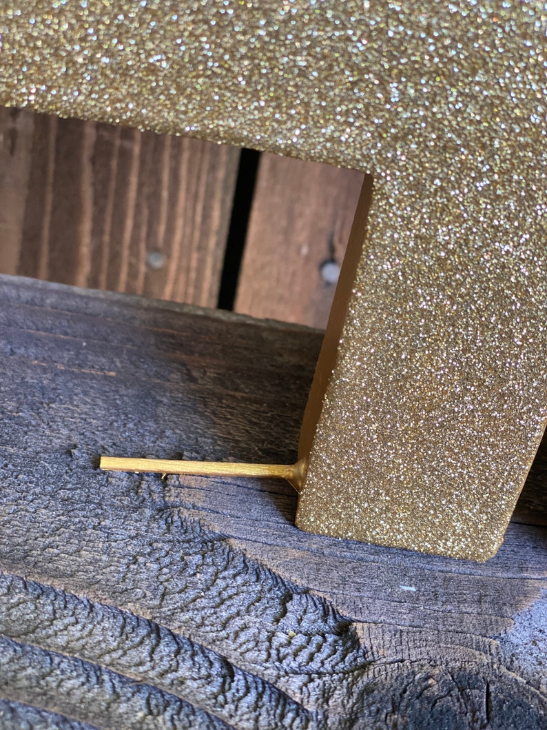 May include: A gold glitter letter with a wooden base. The letter is standing on a wooden surface.