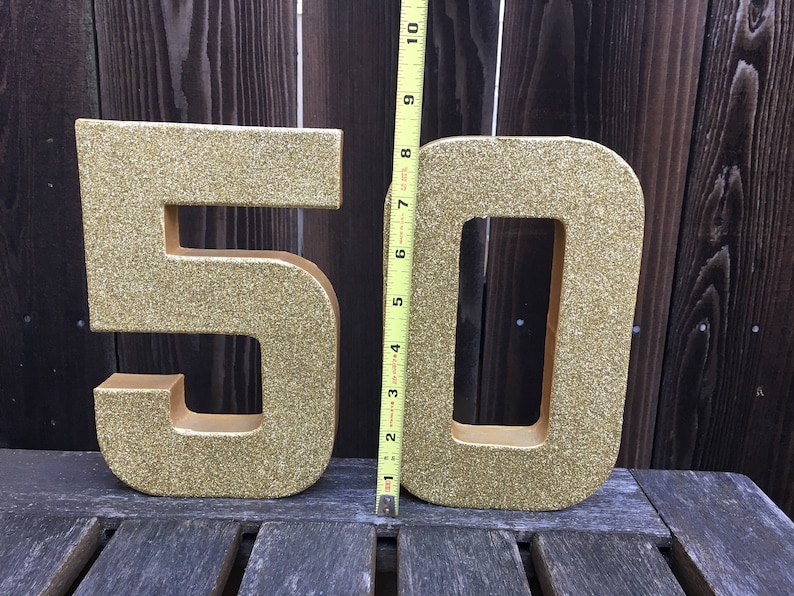 May include: Two gold glitter cardboard numbers, 5 and 0, standing upright on a wooden surface. The numbers are approximately 10 inches tall.