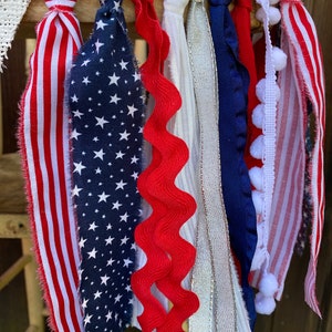 May include: A patriotic bunting with red, white, and blue fabric strips, including a red, white, and blue striped fabric, a blue fabric with white stars, a red wavy fabric, a white fabric with silver glitter, a blue fabric with white trim, a white fabric with white pom-poms, and a red, white, and blue striped fabric with white pom-poms.