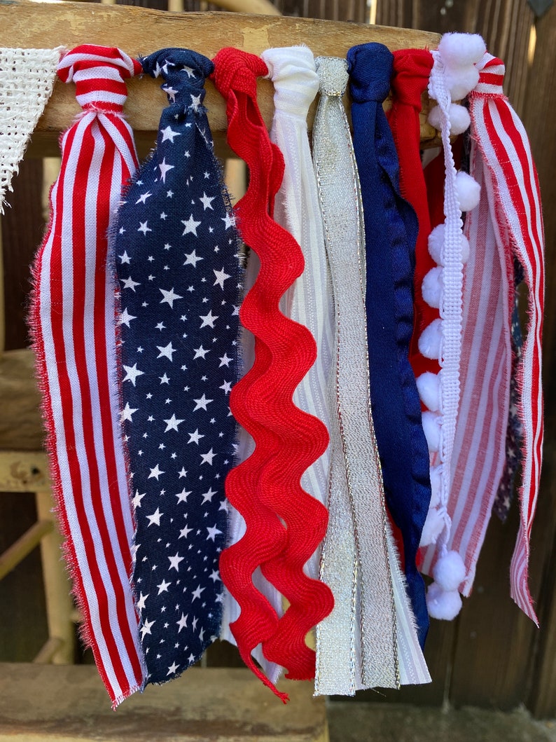May include: A patriotic bunting with red, white, and blue fabric strips, including a red, white, and blue striped fabric, a blue fabric with white stars, a red wavy fabric, a white fabric with silver glitter, a blue fabric with white trim, a white fabric with white pom-poms, and a red, white, and blue striped fabric with white pom-poms.