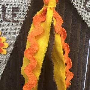 May include: A close-up of a decorative banner with a burlap pennant. The banner features a yellow and orange fabric tassel. The banner is attached to a wooden background. The word "WELCOME" is visible on the banner.