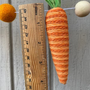May include: A close-up of a carrot-shaped decoration made of orange and white twine. The carrot is about 4 inches long and is hanging from a string. A ruler is visible in the background, showing the length of the carrot.
