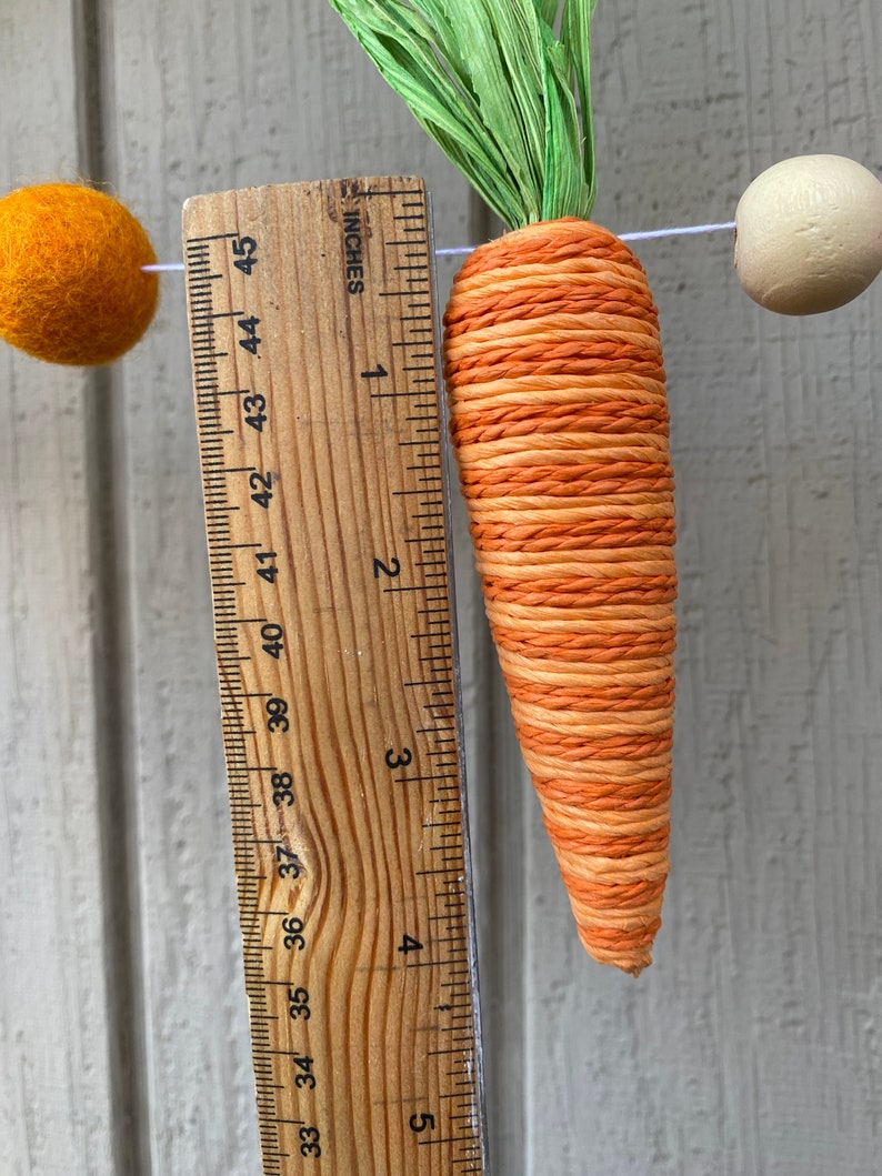 May include: A close-up of a carrot-shaped decoration made of orange and white twine. The carrot is about 4 inches long and is hanging from a string. A ruler is visible in the background, showing the length of the carrot.