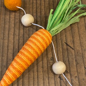 May include: A carrot-shaped garland made of orange and yellow twine with green paper leaves. The garland is strung with wooden beads and orange felt balls.