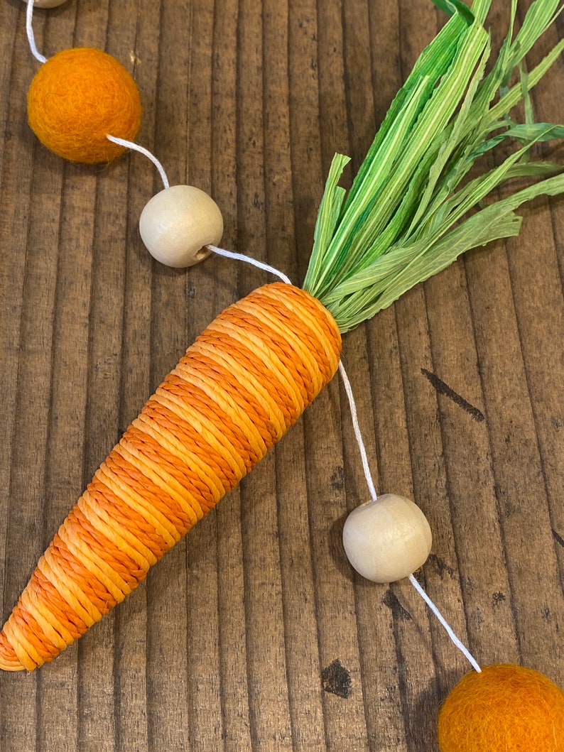 May include: A carrot-shaped garland made of orange and yellow twine with green paper leaves. The garland is strung with wooden beads and orange felt balls.