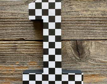 May include: Black and white checkered number one cutout. The number is made of cardboard and has a black border. The background is a wooden surface.