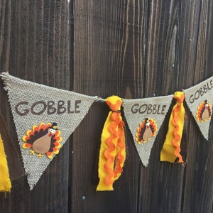 May include: Thanksgiving banner with burlap pennants. Each pennant has the word "GOBBLE" in brown lettering and a turkey applique. The banner is decorated with yellow and orange fabric accents, creating a festive look.