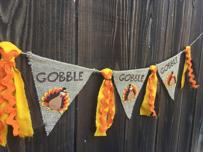 May include: Thanksgiving banner with burlap pennants. Each pennant has the word "GOBBLE" in brown lettering and a turkey applique. The banner is decorated with yellow and orange fabric accents, creating a festive look.