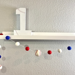 May include: A white shelf with a white number one with red stitching, decorated with a baseball-themed garland. The garland is made of white, red, and blue felt balls.
