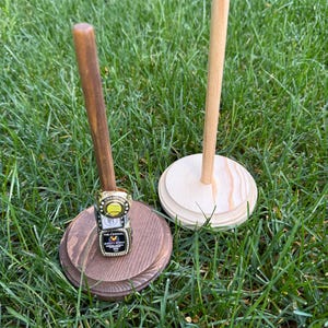 May include: Two wooden display stands with a ring on one. The ring is gold, black, and white, with the words "CHAMPION" and "THE ANNUAL" visible. The stands are on a bed of green grass.