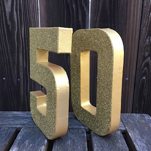 May include: Two large gold glitter foam numbers, 50, standing on a wooden surface. The numbers are three-dimensional and have a sparkly finish.