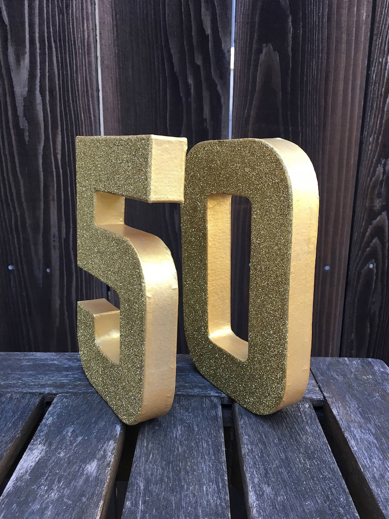 May include: Two large gold glitter foam numbers, 50, standing on a wooden surface. The numbers are three-dimensional and have a sparkly finish.