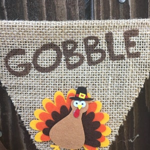 May include: Thanksgiving banner with the word "GOBBLE" in brown letters. A felt turkey with orange, yellow, and brown feathers and a black hat is centered below the word. The banner is made of burlap.