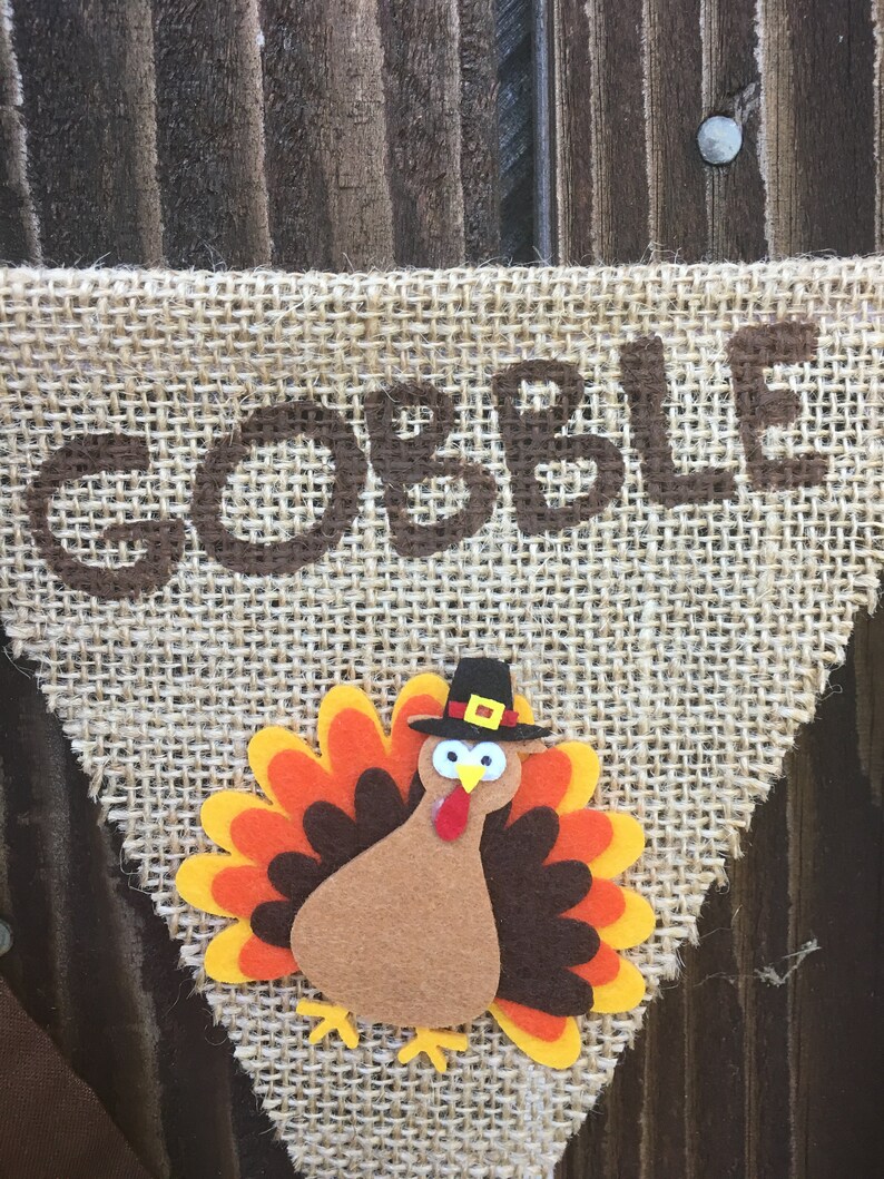 May include: Thanksgiving banner with the word "GOBBLE" in brown letters. A felt turkey with orange, yellow, and brown feathers and a black hat is centered below the word. The banner is made of burlap.