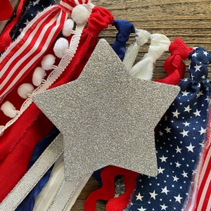 May include: A silver glitter star with red, white, and blue fabric scraps and ribbons arranged around it. The fabric scraps have a variety of patterns, including stripes, stars, and pom-poms.