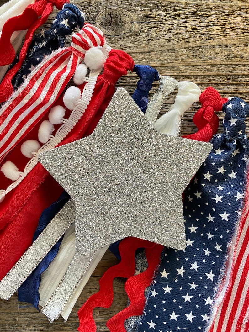 May include: A silver glitter star with red, white, and blue fabric scraps and ribbons arranged around it. The fabric scraps have a variety of patterns, including stripes, stars, and pom-poms.