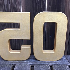 May include: Two large, gold-colored cardboard numbers, 20, standing upright on a wooden surface.
