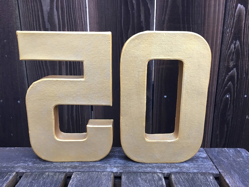 May include: Two large, gold-colored cardboard numbers, 20, standing upright on a wooden surface.