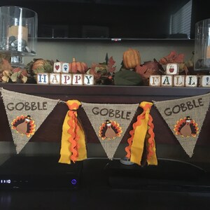 May include: Thanksgiving-themed banner with burlap pennants, each displaying the word "GOBBLE" and a turkey illustration. The banner is adorned with orange and yellow ribbons. Decorative blocks spell "HAPPY FALL!" with pumpkins and leaves.