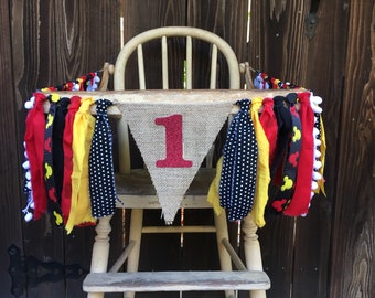 May include: A burlap banner with the number "1" in red glitter, decorated with a black, red, yellow, and white fabric garland. The banner is hanging on a wooden high chair.
