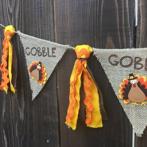 May include: Thanksgiving banner with burlap pennants. Each pennant has the word "GOBBLE" in brown letters and a turkey applique. The banner is decorated with orange and yellow fabric tassels. The background is a dark wood.