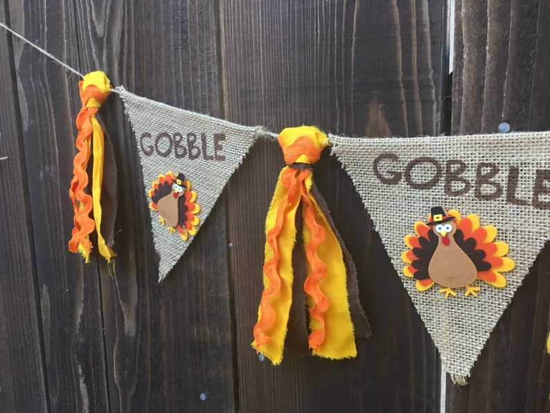 May include: Thanksgiving banner with burlap pennants. Each pennant has the word "GOBBLE" in brown letters and a turkey applique. The banner is decorated with orange and yellow fabric tassels. The background is a dark wood.