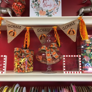 May include: Thanksgiving-themed display with a red background. Features a banner that says "GOBBLE" with turkey designs, glass jars filled with candy, and a sign that says "Give Thanks". Shelves are lined with colorful ribbons.