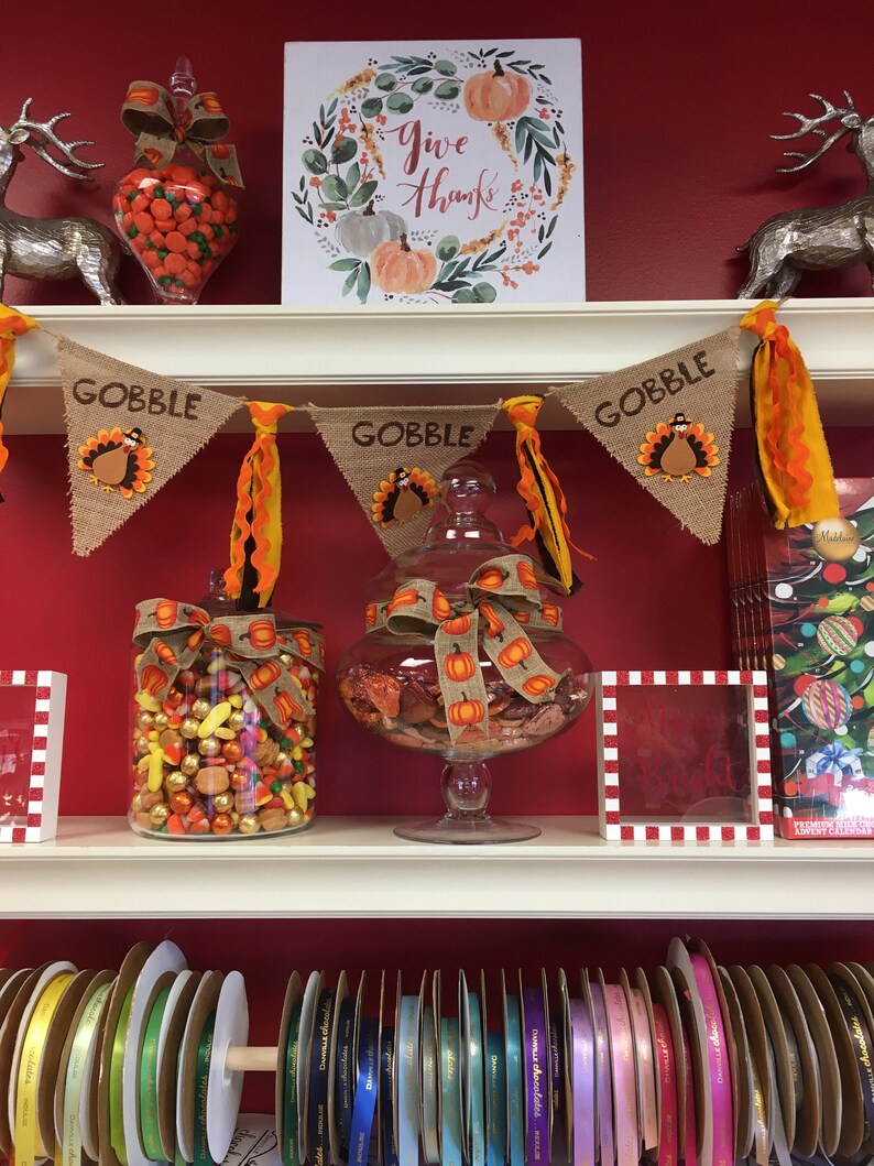 May include: Thanksgiving-themed display with a red background. Features a banner that says "GOBBLE" with turkey designs, glass jars filled with candy, and a sign that says "Give Thanks". Shelves are lined with colorful ribbons.
