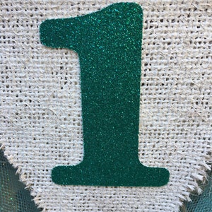 Puede incluir: Un n&uacute;mero uno de purpurina verde sobre un fondo de arpillera blanca.