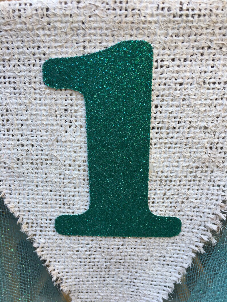 Puede incluir: Un n&uacute;mero uno de purpurina verde sobre un fondo de arpillera blanca.