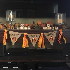May include: Thanksgiving-themed home decor featuring a burlap banner with "GOBBLE" text and turkey illustrations. The display includes pumpkins, fall leaves, and wooden blocks spelling "HAPPY FALL!" with candles in glass holders.