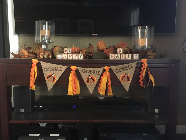 May include: Thanksgiving-themed home decor featuring a burlap banner with "GOBBLE" text and turkey illustrations. The display includes pumpkins, fall leaves, and wooden blocks spelling "HAPPY FALL!" with candles in glass holders.